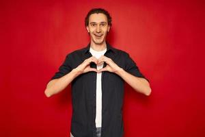 young handsome man dressed in casual clothes, blue shirt isolated on red background, smiles in love, making shape of heart symbol with his hands. concept romance, pride, valentine's day, relationships