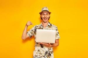 male tourist traveler with laptop in his hands on vacation received message by email with joyful news. he points his finger at device. isolated on yellow background. space for text. concept recreation photo