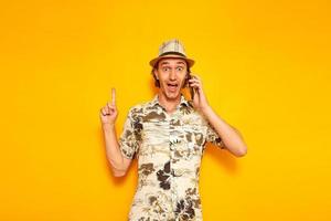 surprised, happy male tourist talking on smartphone points his finger up at an empty area for advertising. isolated on yellow background with space for text. concept of people, vacation, communication photo