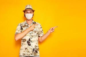 surprised male tourist traveler in medical mask points with his index fingers to side at an empty area for advertising. isolated on yellow background. copy space. concept people, vacation, advertising photo