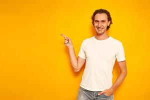 young curly haired smiling man in casual clothes points with his index finger at an empty area for advertising, promotions, discounts. isolated on yellow background with space for text. concept people photo