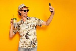 male tourist traveler in hat, Hawaiian shirt, sunglasses on vacation with drink in cocktail glass takes selfie on phone, communicates via video link. isolated on yellow background with space for text photo