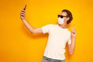 Man takes selfie, makes video call, points at camera of smartphone, enjoys spare time, dressed in casual clothes and sunglasses, medical mask shows peaceful gesture, isolated on yellow background photo