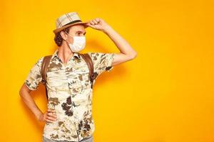 a surprised male tourist traveler with a backpack in a medical mask holds a hat with his hand and looks away. isolated on a yellow background with space for text. concept of people, travel, vacation photo