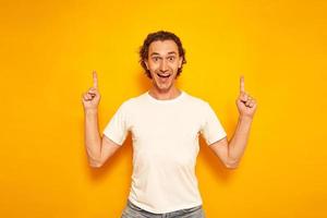 an excited curly-haired man in casual clothes looks forward, points up with his index fingers at an empty area for advertising, attracts attention. isolated on a yellow background with space for text. photo