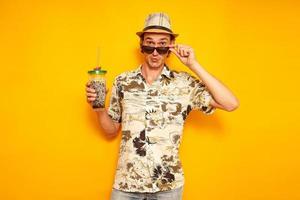 pleasantly surprised face, male tourist traveler in hat, Hawaiian shirt on vacation with drink in cocktail glass lowered his sunglasses with his hand. isolated on yellow background with space for text