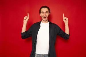 an excited, joyful, smiling man in casual clothes points with his index fingers at an empty free place for advertising text. isolated on red background. christmas promotion, discounts, copy space, buy photo