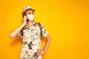 A happy male tourist traveler in a medical mask with a phone in his hand is talking on a smartphone. isolated on a yellow background with space for text. concept of people, vacation, communication photo