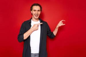 isolated shot of contented European young man making small gesture with hand measuring tiny object, with smiling expression on face dressed in casual clothes, dark blue shirt, demonstrates small thing photo