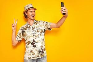 happy male tourist traveler with phone in his hand takes selfie, communicates via video communication on voyage on vacation. isolated on yellow background. concept - people, technology, communication photo