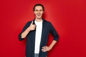 joyful happy smiling man in blue shirt, white T-shirt holds hand in front of him and shows his thumb. isolated on red studio background with space for text. concept - approval, recommendation, people photo