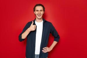 joyful happy smiling man in blue shirt, white T-shirt holds hand in front of him and shows his thumb. isolated on red studio background with space for text. concept - approval, recommendation, people photo