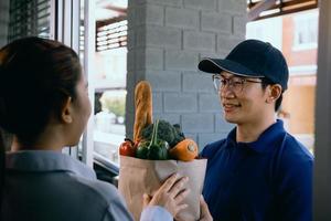 Delivery asian man handing a paper bag inside with vegetables or food to an Asian woman in front of his house. photo