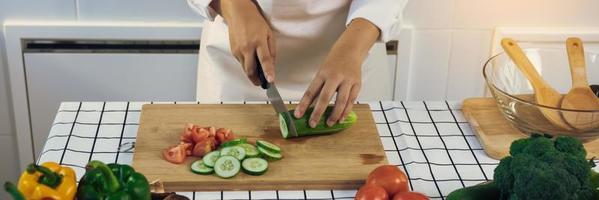 Woman using knife and hands cutting cucumber on wooden board in kitchen room. photo