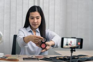 Young asian woman vlogging about cosmetics skin care items products on table with her video camera and demonstrates product use and reviews for her online blog channel.