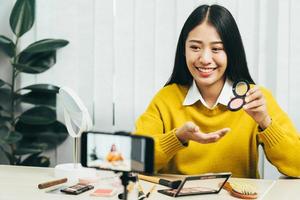 una joven asiática que hace vlogs sobre productos cosméticos para el cuidado de la piel en la mesa con su cámara de video y demuestra el uso de productos y reseñas para su canal de blog en línea.