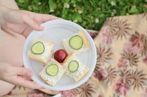 a young girl holds a plate with four small sandwiches with melted cheese ,cucumber slices and one plum in the center..Healthy summer picnic in the Park or in the garden. photo