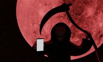 el cráneo satánico con un sombrero negro con capucha y una hoz es un arma. el fondo de la luna llena roja en la noche de la representación 3d muerta foto