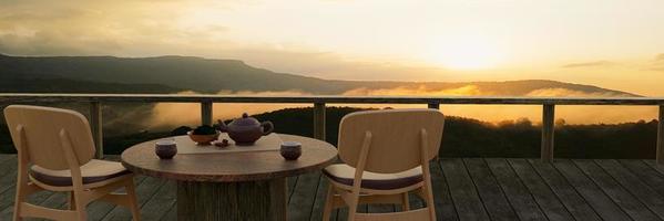 juego de té de cerámica y hojas de té secas sobre una mesa y silla de madera en el balcón o terraza de madera. paisaje de montaña con niebla matutina y sol. té caliente en el ambiente de montaña. representación 3d foto