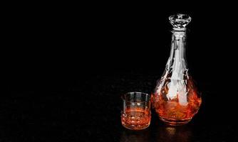 The bottle and glass have an elegant pattern for brandy or whiskey. The glass bottle has a diamond-shaped cork. The bottle and the glass are crystal-patterned with a black background and a black photo