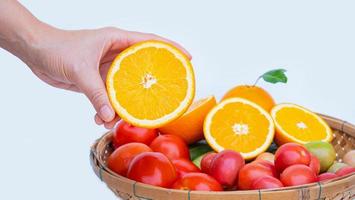 la mano izquierda femenina sostiene una naranja medio madura en la mano. cesta de bambú poner verduras y frutas ricas en vitamina c como naranjas, tomates sobre fondo blanco. foto