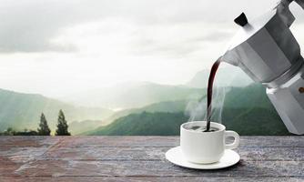 vierta café recién hecho o espresso de la cafetera moka en una taza de café con leche. café caliente en una taza colocada sobre la mesa o balcón de madera. vista a la montaña por la mañana, sol por la mañana. representación 3d foto