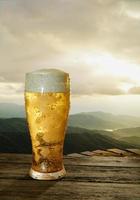 vaso de cerveza transparente con cerveza fría y espuma en la boca del vaso y las gotas de agua se adhieren a los bordes. tablón o superficie de madera y con paisaje de montaña bajo el sol de la mañana. representación 3d foto