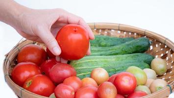 Female left hand Hold the tomato in your hand. Bamboo basket put available fruits and vegetables like cucumber and tomato on white background. photo