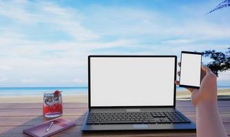 A computer or laptop placed on a wooden table, a smartphone in the hands of a person. The screen is blank white. Sea and beach background. Work Out, Beach Vacation, 3D Rendering photo