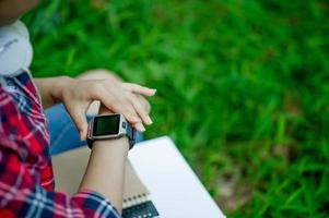 The girl watches the watch in hand, watches the time in a black watch, wears a red shirt and a green background. And there is a copy space. photo