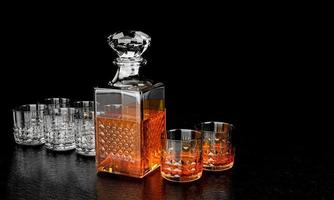 The bottle and glass have an elegant pattern for brandy or whiskey. The glass bottle has a diamond-shaped cork. The bottle and the glass are crystal-patterned with a black background and a black photo