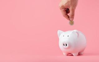 Money saving and investment concept. Hand putting coin to piggy bank on pink background with copy space photo