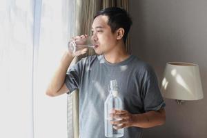 Asian Man drinking mineral water and looking outside through windows bedroom photo