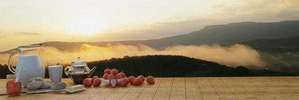 desayuno panorámico café con leche fresca en un vaso y una cafetera. pan de mantequilla en una tabla de cortar de madera. muchas fresas frescas. montaña por la mañana con niebla y el sol brilla. representación 3d foto