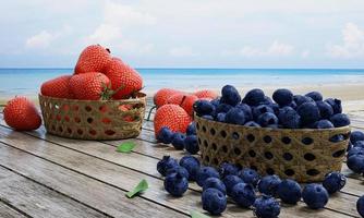 fresas y arándanos frescos en una cesta de bambú. baya sobre una superficie de madera. el paisaje de fondo es el mar y la playa, el cielo azul y las nubes blancas, el paisaje y la luz del sol por la mañana. representación 3d foto