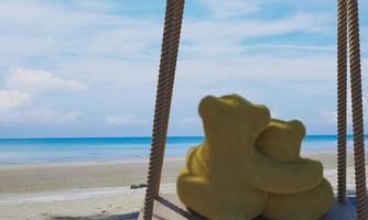 The brown teddy bear hug, hugging each other with love and romance. Lovers sit on a wooden swing tied with a rope by the beach, sea, bright blue sky, and white clouds. 3D Rendering photo