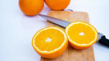 Ripe oranges are cut in half and placed on a wooden cutting board. With a sharp knife placed on the ground and a white background photo