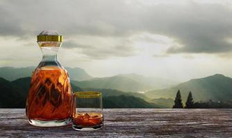 whisky o brandy en una botella transparente con diseño en espiral y un vaso transparente colocado sobre corteza de árbol o corteza de árbol. paisaje con pinos y montañas por la mañana. está amaneciendo. representación 3d foto