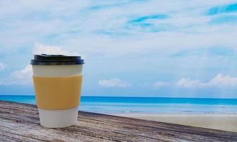 Plastic coffee cup with black plastic mat and lid Place on a wooden floor table. Turquoise sea and sandy beach The sky is bright with white clouds. 3D Rendering photo