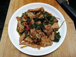 revuelva la carne frita con albahaca sagrada en un plato blanco, colocado sobre una tabla de cortar de madera y un cuchillo. la comida popular en tailandia consiste en carne, ajo, chiles frescos y hojas de albahaca. foto