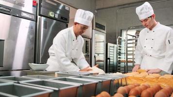 dois chefs asiáticos profissionais em uniformes e aventais de cozinheiro branco estão amassando massa e ovos, preparando pão e alimentos frescos de padaria, assando no forno na cozinha de aço inoxidável do restaurante. video