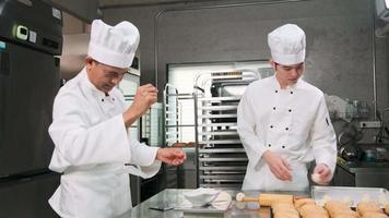 dos chefs asiáticos profesionales con uniformes y delantales de cocinero blanco están amasando masa de pastelería y huevos, preparando pan y comida fresca de panadería, horneando en el horno en la cocina de acero inoxidable del restaurante. video