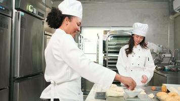 due chef professionisti in uniforme bianca da cuoco e grembiuli impastano la pasta sfoglia e le uova, preparano pane, biscotti e cibi da forno freschi, cuociono in forno in una cucina in acciaio inossidabile di un ristorante. video