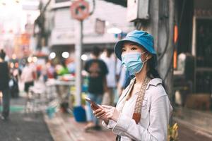 una joven viajera asiática adulta usa mascarilla y usa teléfono móvil. foto