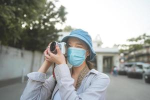 Una joven adulta asiática que viaja con mochila usa mascarilla para tomar una foto con la cámara.