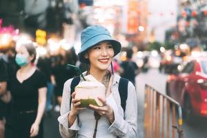 joven adulta asiática gourmet mujer mochila viajero comiendo jugo de coco en la comida callejera de la ciudad china. foto