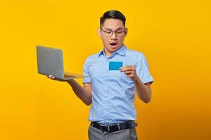 sorprendido joven asiático asiático con anteojos sosteniendo una laptop y mirando una tarjeta de crédito aislada en un fondo amarillo. concepto de empresario y emprendedor