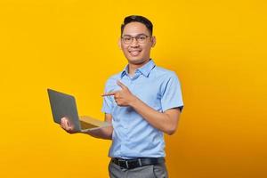 retrato de un alegre y apuesto hombre asiático con anteojos apuntando a la laptop, mostrando algo en línea aislado en un fondo amarillo. concepto de empresario y emprendedor foto