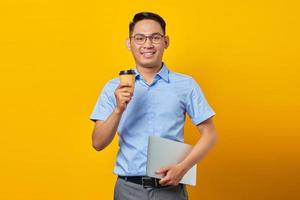 Portrait of smiling handsome Asian man in glasses holding laptop and showing cup coffee isolated on yellow background. businessman and entrepreneur concept photo