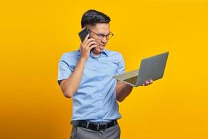 Angry young asian man Asian in glasses talking on mobile phone and using laptop isolated on yellow background. businessman and entrepreneur concept photo